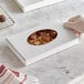 A hand holding a white candy box with an oval window containing chocolates.