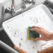 A hand cleaning a white Baker's Mark aluminum sheet pan with a sponge.