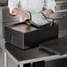 A chef using a Vollrath black food pan carrier to put food in a metal container.