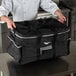 A chef in a white coat holding a large black Vollrath food pan carrier bag.