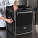 A man in a chef's uniform holding a large black Vollrath 5-Series insulated tower bag.
