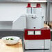 A Robot Coupe R2N food processor on a counter next to a stainless steel bowl of cucumbers.