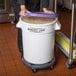 A woman standing in front of a large white Baker's Lane ingredient storage bin with a purple lid and gray dolly in a school kitchen.