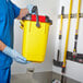 A person wearing blue scrubs holding a yellow Rubbermaid bucket.