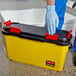 A hand in a blue glove holding a yellow and black Rubbermaid box.