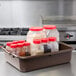 A Tablecraft brown plastic bus tub on a counter filled with plastic containers with red lids containing various spices.