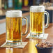 Two Libbey paneled beer mugs filled with beer on a table with a bowl of nuts.