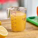 A purple Choice 1 Cup plastic measuring cup with yellow liquid next to a lemon slice.