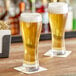 Two Acopa pilsner glasses of beer on a table.