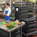 A woman in a blue apron using a black Metro Mightylite front loading food pan carrier on a cart.