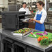 A woman in a blue apron opening a black Metro Mightylite front loading food pan carrier on a counter.