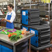 A woman in a blue apron using a Metro Mightylite black and blue food pan carrier.