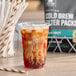 A plastic cup filled with iced cold brew coffee with cream swirling inside, placed on a wooden surface next to a striped paper straw and a bag labeled 'Cold Brew Filter Packs.'