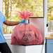 A person in blue scrubs and gloves putting a red plastic bag in a trash can.