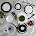 A white Arcoroc round rimless glass plate on a table with fruit, leaves, a spoon, and a glass.