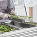 A person in a white uniform using a Vollrath long stainless steel food pan to prepare food on a counter in a salad bar.