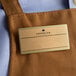 A brown apron with a customizable gold rectangle name tag on it.
