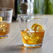 Two Libbey plastic rocks glasses with whiskey and ice on a table with a lemon peel.