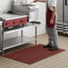 A man in a chef's uniform standing on a red rubber anti-fatigue floor mat in a professional kitchen.