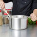 A person in a black coat stirring food in a Vollrath stainless steel sauce pot.