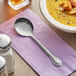 A Choice stainless steel bouillon spoon on a napkin next to a bowl of soup.