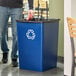 A person putting a can of soda in a blue Rubbermaid Untouchable recycling bin.