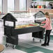 A black 6-foot portable youth food and salad bar with an open base and clear sneeze guard, shown in a cafeteria setting.