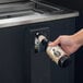 A hand using an Avantco bottle opener to open a beer bottle over a bottle cap catcher on a counter.