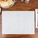 A table with a white Dubonnet placemat and silverware with a bowl of bread on a wooden table.