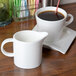 An Arcoroc Daring white porcelain creamer on a table with a cup of coffee.