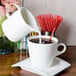 A person pouring milk into a white cup of coffee on a green coaster using an Arcoroc porcelain creamer.