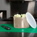 A beige Cambro round plastic crock with lid on a cutting board with broccoli and a knife.