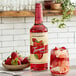 A Torani Puremade Strawberry Flavoring Syrup bottle next to a glass of ice and strawberries.
