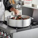 A person cooking meat in a Choice aluminum brazier on a stove.