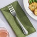 A Libbey stainless steel dinner fork on a green napkin next to a plate of food.