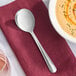 A Libbey Windsor stainless steel round bowl soup spoon on a red napkin next to a bowl of soup.