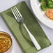 A Libbey stainless steel dinner fork on a green napkin next to a plate of broccoli.