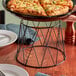 An American Metalcraft black metal wire frame pizza stand holding a plate of pizza on a table.