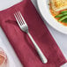 A Libbey Windsor stainless steel dinner fork on a red napkin next to a plate of food.