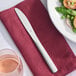 A Libbey Windsor Grandeur stainless steel dinner knife on a red napkin next to a plate of food.
