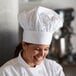 A woman wearing a white Mercer Culinary chef's hat smiles in a professional kitchen.