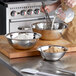 A hand holding a whisk mixing ingredients in a Vollrath stainless steel mixing bowl on a table.