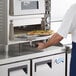 A man using an Avantco countertop pizza oven to cook a pizza on a square pizza stone.