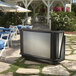 A black and silver Cambro portable bar set up outside with a blue tablecloth on a table and white chairs.