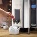 A person using a Waring hot water boiler to fill a white teapot with water.