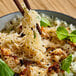 A bowl of cooked rice noodles garnished with herbs and spices, being lifted with chopsticks.