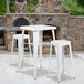 A white metal Flash Furniture bar height table with 2 square stools on an outdoor patio next to a stone fireplace.