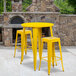 A yellow metal table and two square yellow metal bar stools on a stone patio.