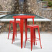 A red metal round bar table with red metal legs and two square backless stools with red metal legs.