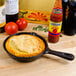 An American Metalcraft pre-seasoned mini cast iron skillet with cornbread next to a bottle of hot sauce.